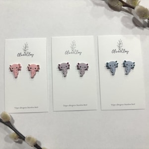Puede incluir: Tres pares de pendientes de botón de axolotl en rosa, morado y azul, expuestos en tarjetas blancas. Las tarjetas tienen el texto "Olives&Clay" y "Hypo-allergenic stainless steel". Los pendientes son pequeños y detallados.