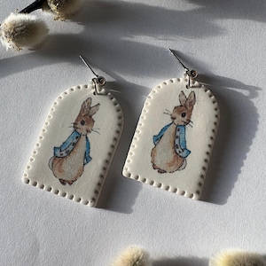 May include: A pair of white ceramic earrings featuring a hand-painted illustration of Peter Rabbit wearing a blue jacket. The earrings have a rectangular shape with rounded top edges and a dotted border. Silver-colored ear wires are attached to the top of each earring.