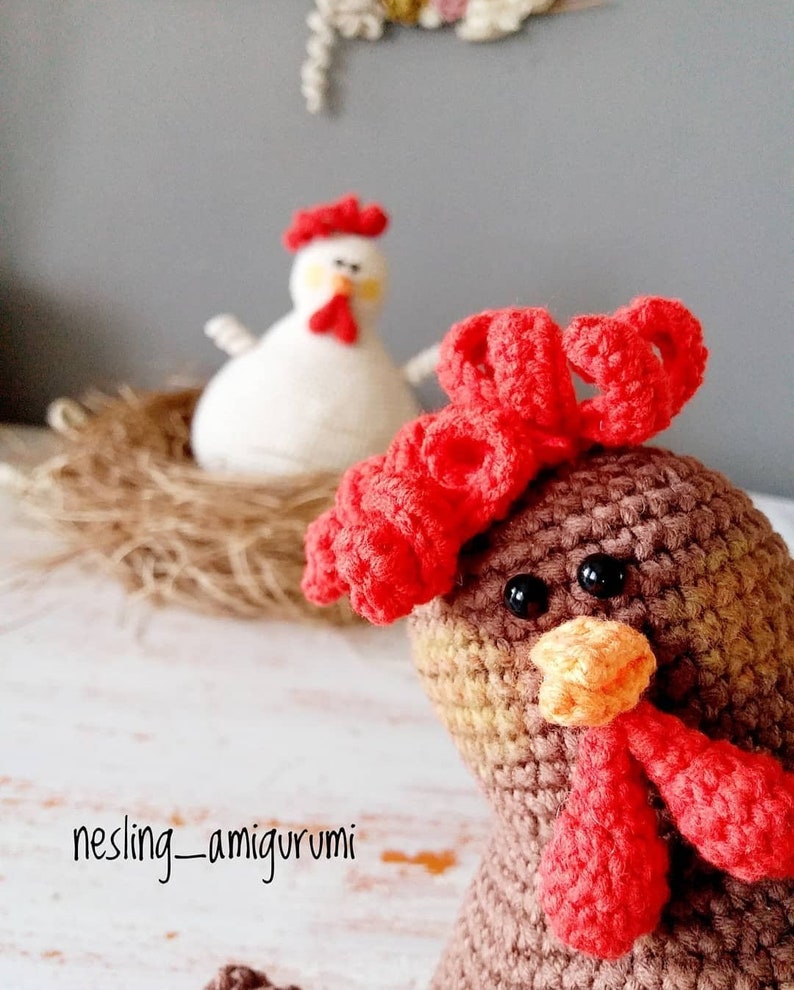 May include: Two crocheted chickens, one brown and one white, with red combs and wattles. The brown chicken has a red bow on its head. The chickens are sitting on a bed of straw.