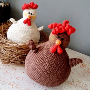 May include: Two crocheted chicken toys, one white and one brown, with red combs and beaks. The white chicken is sitting in a nest of straw, while the brown chicken is on a white wooden surface.