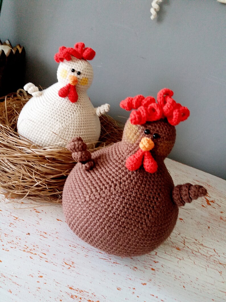 May include: Two crocheted chicken toys, one white and one brown, with red combs and beaks. The white chicken is sitting in a nest of straw, while the brown chicken is on a white wooden surface.