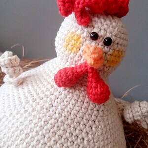 May include: A crocheted white chicken toy with a red comb, orange beak, and yellow spots on its face. The chicken is sitting in a nest of brown straw.