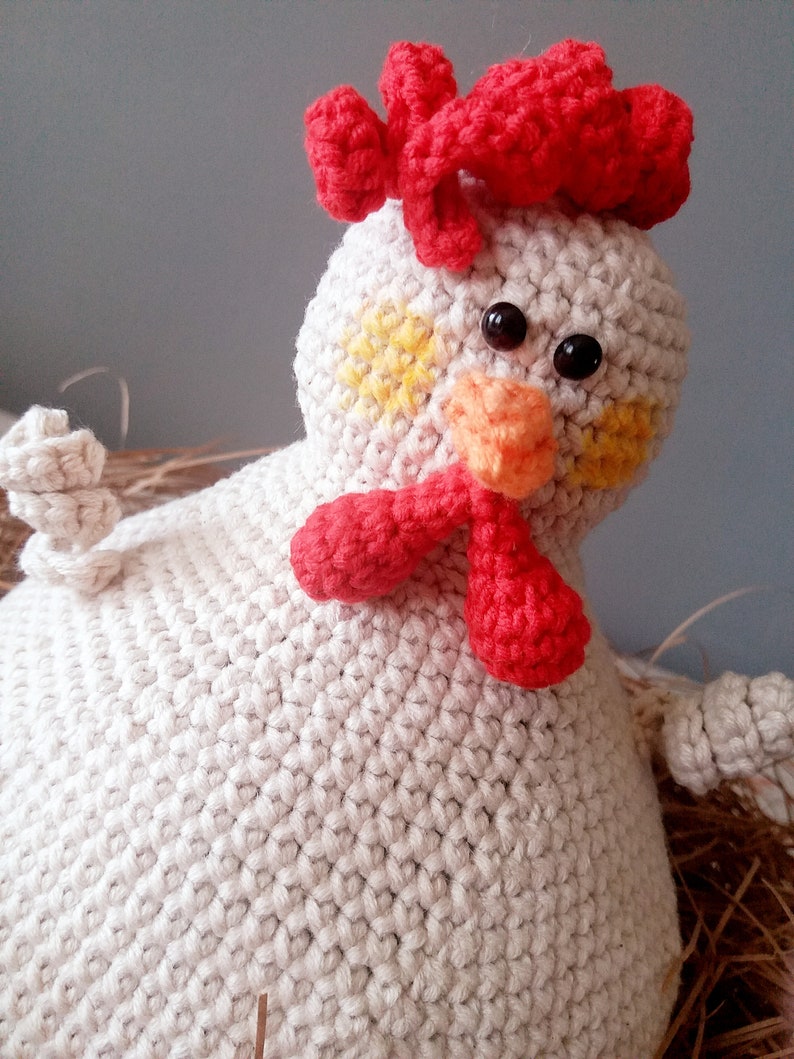 May include: A crocheted white chicken toy with a red comb, orange beak, and yellow spots on its face. The chicken is sitting in a nest of brown straw.