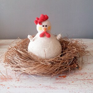 May include: A white crocheted chicken with a red comb and yellow eyes sits in a brown nest made of straw. The chicken is sitting on a white wooden surface.