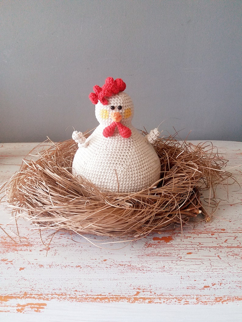 May include: A white crocheted chicken with a red comb and yellow eyes sits in a brown nest made of straw. The chicken is sitting on a white wooden surface.