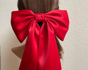 May include: A large, vibrant red hair bow crafted from a smooth, satin-like fabric. The accessory features a central knot with two wide, structured loops and two long, flowing tails that extend downward, creating a classic and bold hair ornament.