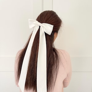 Op de afbeelding: Een wit satijnen lint haar strik met lange staarten. De strik is in een eenvoudige knoop geknoopt en de staarten zijn aan de onderkant met een kleine knoop aan elkaar geknoopt. Het lint is geborduurd met de letters "P.K."