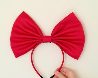 May include: A vibrant red headband featuring a large bow. The bow is made of a smooth, possibly satin-like fabric, and is attached to a thin, red headband. The ends of the headband are finished with black tips.