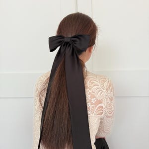 May include: A black satin ribbon tied into a large bow, worn as a hair accessory. The ribbon is long and flows down the back of the person's head.