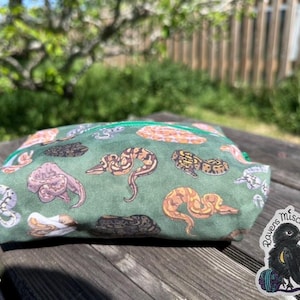 May include: A green zipped pouch with a repeating pattern of snake illustrations in brown, tan, and purple. The pouch is on a wooden surface with a sticker of a raven.