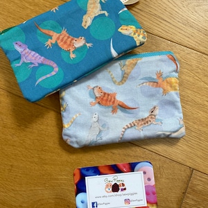 May include: Two small zippered pouches with a blue and white background featuring a repeating pattern of colourful bearded dragons. The pouches are handmade and have a tag that says "Handmade". A business card with the text "Sew Piggies" and a website address is also in the image.