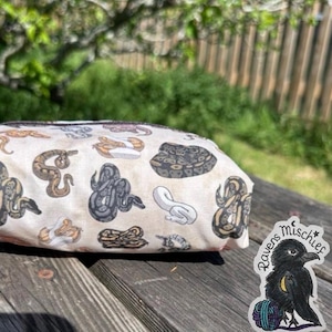May include: A rectangular, cream-colored zippered pouch featuring a repeating pattern of various snake illustrations in shades of brown, gray, and white. A sticker with a raven and the text "Ravens Mischief" is on the right.