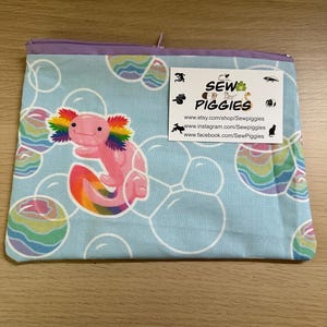 May include: A light blue zippered pouch with a rainbow axolotl design and a white background. The pouch has a repeating pattern of rainbow bubbles. The pouch has a purple zipper and a white tag with the text "Sew Piggies" and the website address "www.etsy.com/shop/Sewpiggies".