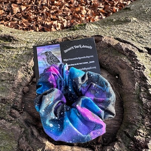 May include: A fabric scrunchie with a galaxy print in shades of blue, purple, and black, with white speckles. The scrunchie is resting on a textured, brown tree trunk. A business card with the text "Raven's Dark Jewels" is also visible.