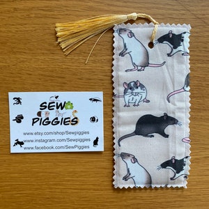 May include: A beige fabric bookmark with a white tassel. The bookmark features a repeating pattern of black and white rats. The bookmark has a scalloped edge and a hole punched in the top corner. The bookmark is handmade and has the text "Sew Piggies" on it.