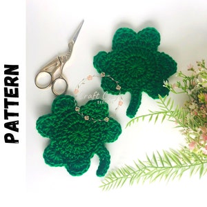 Peut inclure: Deux trèfles à quatre feuilles en crochet vert sur un fond blanc. Les trèfles sont faits avec un fil vert et ont une tige. Il y a une paire de ciseaux dorés et un bouquet de fleurs roses en arrière-plan.