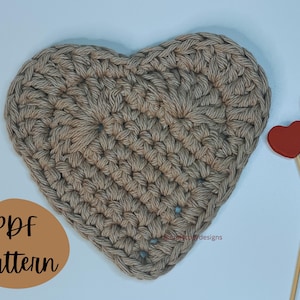 Op de afbeelding: Een gehaakte hartvormige onderzetter in beige kleur. De onderzetter ligt op een wit oppervlak met twee rode houten harten op stokjes.