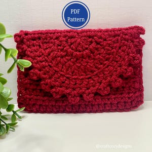 May include: A handmade, red crochet clutch with a textured pattern and scalloped flap. The image includes a blue circle with the text "PDF Pattern". The clutch is a deep red color and appears to be made of soft yarn.