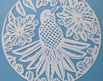 May include: A white paper cut-out of a bird surrounded by flowers and leaves, set against a light blue background. The bird has a detailed, geometric pattern on its body. The design is circular, with intricate details.