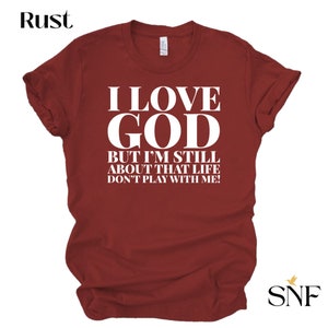 May include: A maroon t-shirt with white text that reads "I LOVE GOD BUT I'M STILL ABOUT THAT LIFE DON'T PLAY WITH ME!"