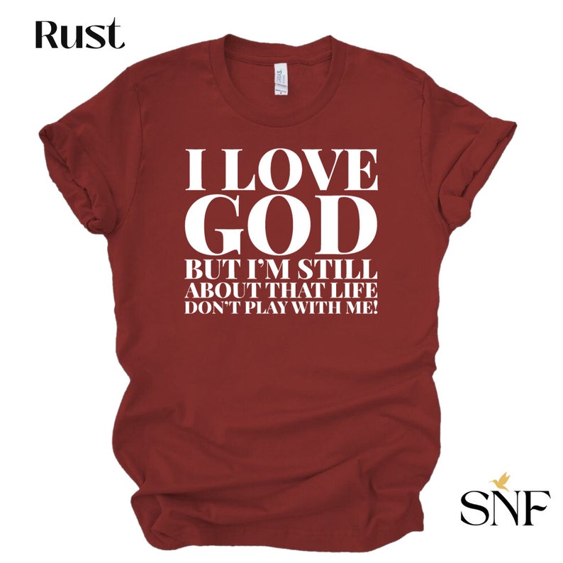 May include: A maroon t-shirt with white text that reads "I LOVE GOD BUT I'M STILL ABOUT THAT LIFE DON'T PLAY WITH ME!"
