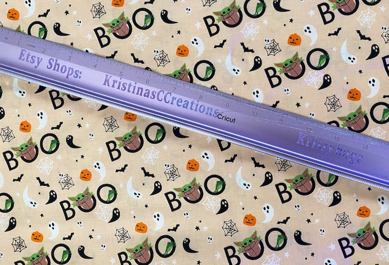 May include: A beige fabric with a repeating pattern of black "BOO" text, green and brown Baby Yoda characters, orange pumpkins, white ghosts, black bats, and white spiderwebs.