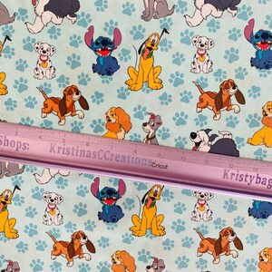 Op de afbeelding: Lichtblauwe stof met cartoonhonden zoals Pluto, Stitch en Dalmatische puppy's, en pootafdrukken. Een roze liniaal met tekst loopt over de afbeelding. Geschikt voor knutsel- of naaiprojecten.
