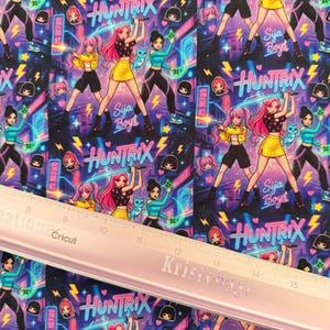 Peut inclure: Tissu à motifs avec des personnages de style anime en poses d'action, tenant des épées lumineuses. Le motif comprend le texte "Huntrix" et "Sija Boys" dans une police de style néon, sur fond violet foncé.