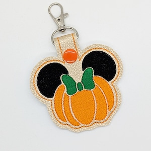 May include: A beige leather keychain with a pumpkin-shaped design. The pumpkin is orange with a green stem and a green bow. The keychain has black mouse ears and a red button.