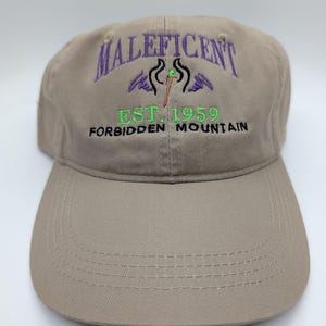 May include: A tan baseball cap with the word "Maleficent" embroidered in purple, along with a graphic and the text "EST. 1959 FORBIDDEN MOUNTAIN" in green and black. The cap has a curved brim and a structured crown.