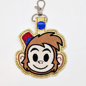May include: A brown monkey face keychain with a red and blue hat, gold glitter trim, and a silver clip.