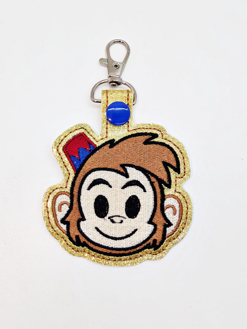 May include: A brown monkey face keychain with a red and blue hat, gold glitter trim, and a silver clip.