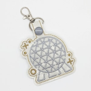 May include: A silver and white keychain featuring a geometric sphere design. The keychain has a silver clasp and a small, gray button. The sphere design is outlined in silver and has a grid-like pattern. The edges are trimmed with gold stitching.