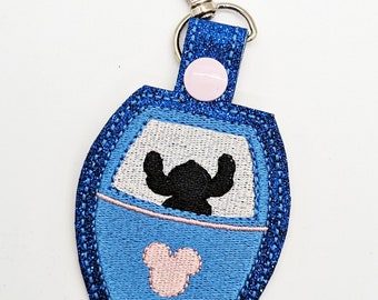 May include: Blue and pink glitter keychain featuring Stitch from Lilo & Stitch in a spaceship with a Mickey Mouse head on the bottom.