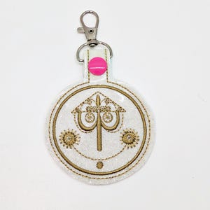 May include: A white glitter keychain with a gold embroidered design of a lamppost with three lights and two suns. The keychain has a pink button and a silver metal clasp.