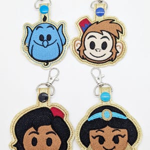 May include: Four embroidered keychains featuring characters from the Disney movie Aladdin. The keychains are gold with glitter and feature Abu the monkey, Jasmine, Aladdin, and Genie.