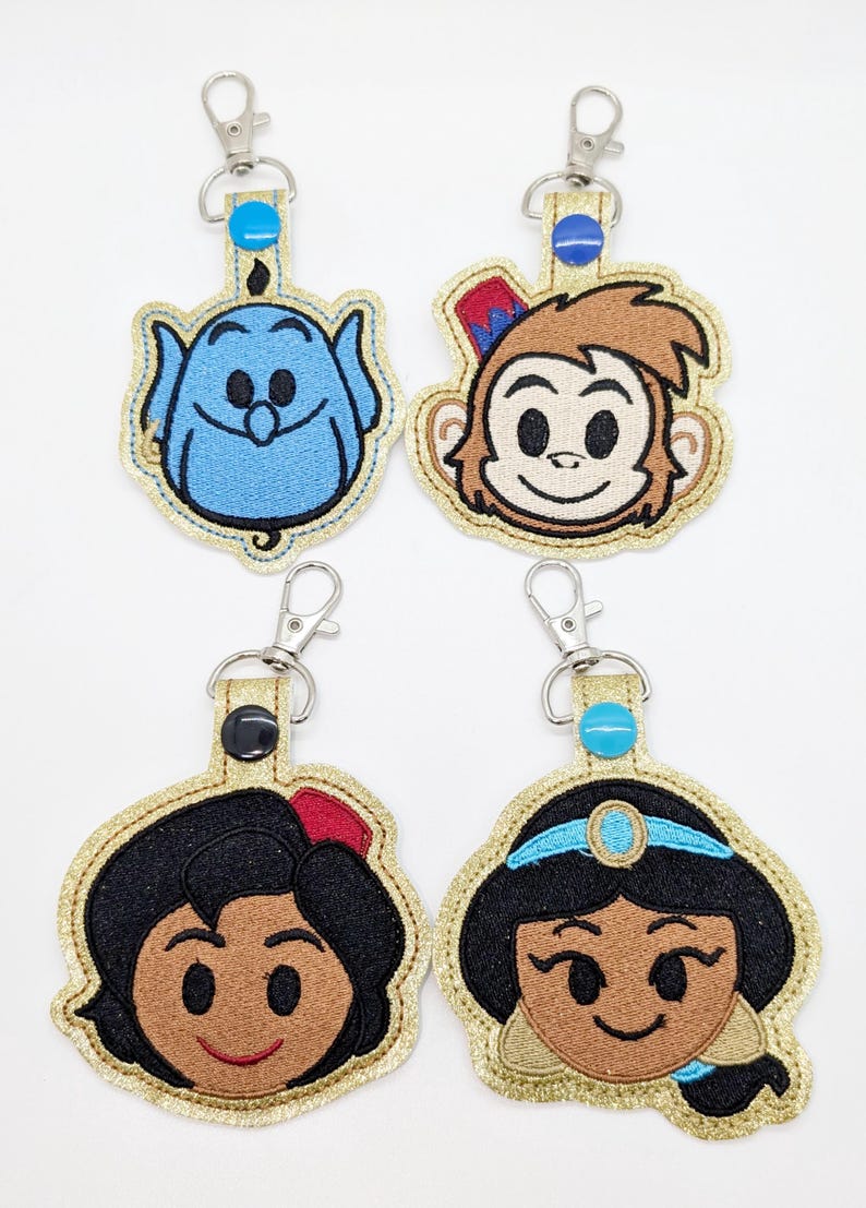 May include: Four embroidered keychains featuring characters from the Disney movie Aladdin. The keychains are gold with glitter and feature Abu the monkey, Jasmine, Aladdin, and Genie.