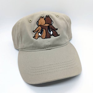 May include: A tan baseball cap featuring an embroidered design of two cartoon chipmunks hugging. The chipmunks are brown and dark brown with white accents. The cap has a curved brim and a classic six-panel design.