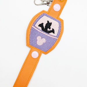 May include: A purple and orange key chain with a white button and a Mickey Mouse silhouette in a ride car. The keychain has a silver metal clasp.