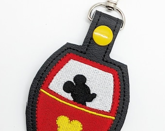 May include: A red and yellow embroidered keychain featuring Mickey Mouse in a ride vehicle. The keychain is attached to a black leather strap with a silver metal clasp.