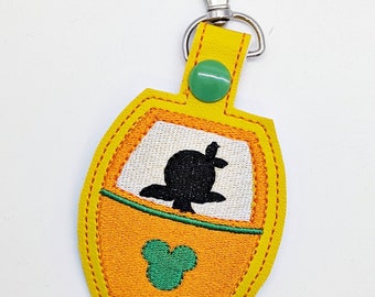 May include: Yellow and green embroidered keychain featuring a silhouette of Mickey Mouse in a ride vehicle. The keychain has a silver clasp.