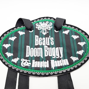 May include: A black and green oval-shaped name tag with white embroidery. The tag reads "Beau's Doom Buggy The Haunted Mansion". The tag has black straps attached to the top.