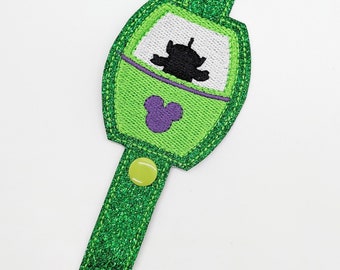 May include: Green and white keychain with a cartoon alien inside a green and white frame. The keychain has a purple Mickey Mouse head and a green glitter strap.