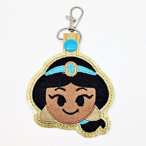 May include: A gold glitter keychain with a blue and black embroidered image of Princess Jasmine from Aladdin. The keychain has a silver metal clasp.