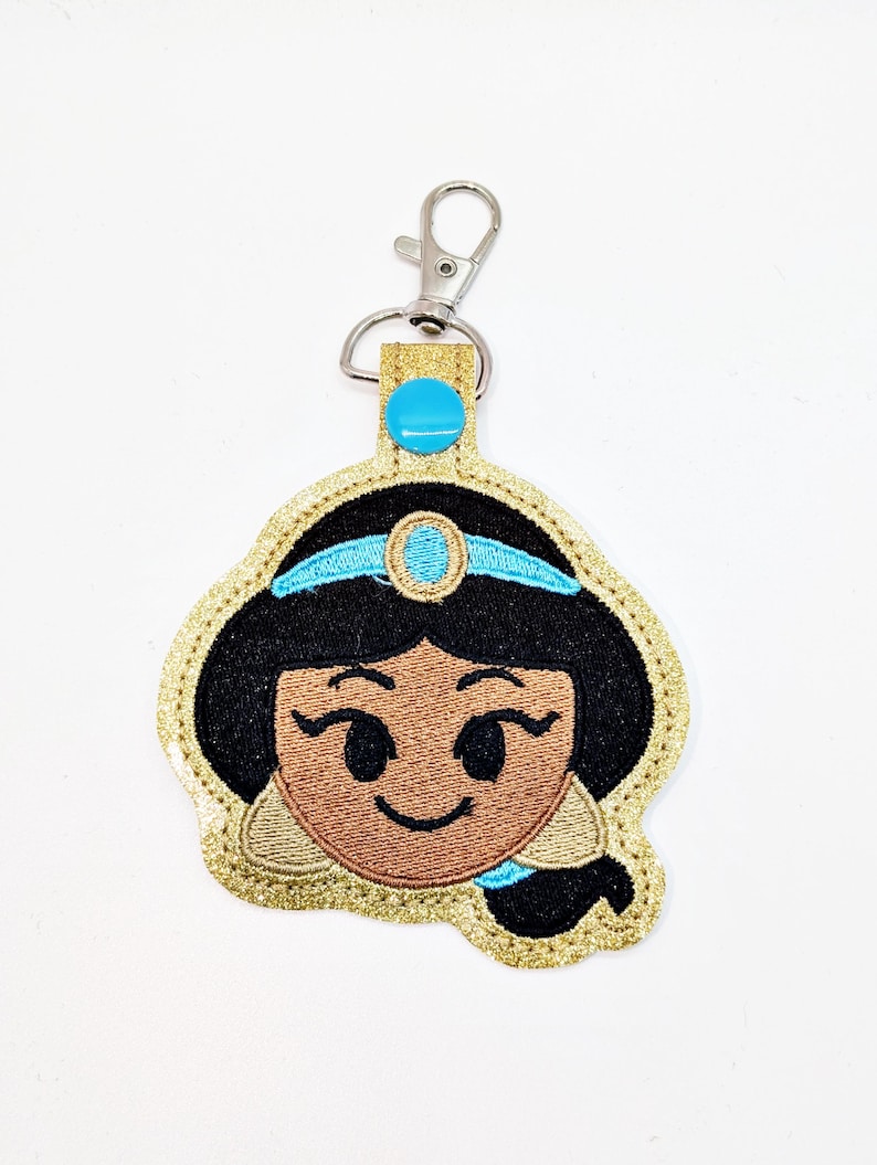 May include: A gold glitter keychain with a blue and black embroidered image of Princess Jasmine from Aladdin. The keychain has a silver metal clasp.