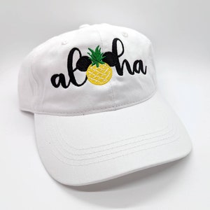 May include: White baseball cap with black embroidered text that says "Aloha" and a yellow pineapple with green leaves.