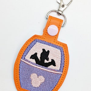 May include: Orange and purple fabric keychain with a silhouette of Mickey Mouse and a silhouette of a ride vehicle with three figures inside. The keychain has a silver metal clasp.