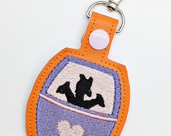May include: Orange and purple fabric keychain with a silhouette of Mickey Mouse and a silhouette of a ride vehicle with three figures inside. The keychain has a silver metal clasp.