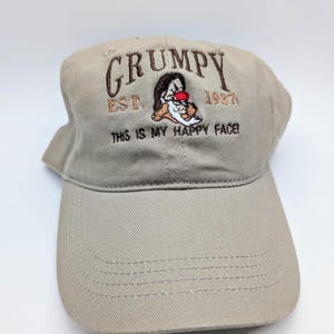 May include: A beige baseball cap with an embroidered image of Grumpy from Snow White and the Seven Dwarfs. The text "GRUMPY EST. 1937 THIS IS MY HAPPY FACE!" is embroidered on the front of the cap.