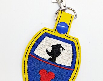 May include: Yellow fabric keychain with a blue and white embroidered image of Mickey Mouse in a ride vehicle. The keychain has a silver clip.